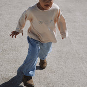 Child wearing a beige long-sleeve shirt and blue pants walking on a concrete surface.