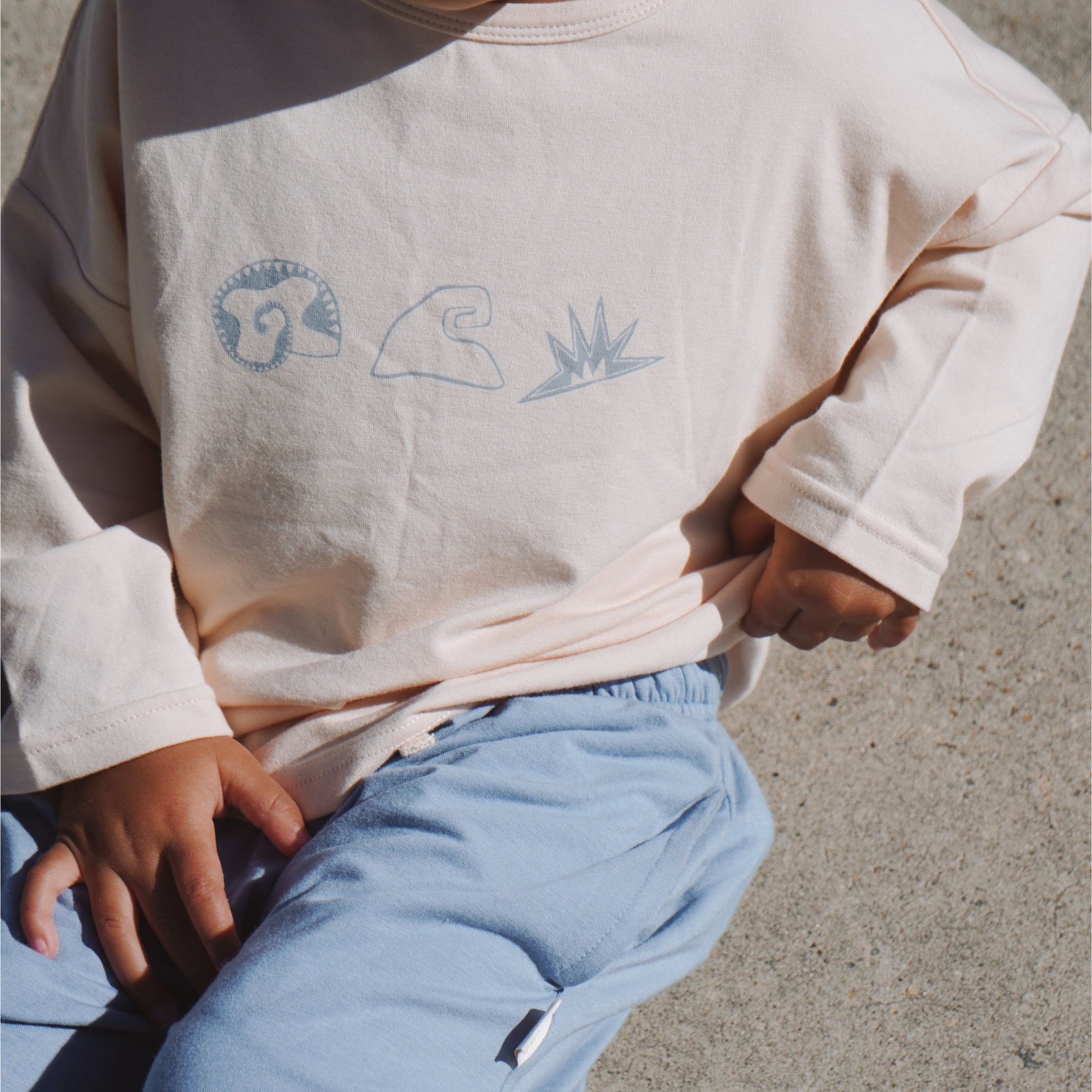 Front view close-up of Beachcomber tee showing minimal shell, wave, and sun motifs on cream fabric.