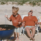 child wearing rust tee on an beach playing with BBQ