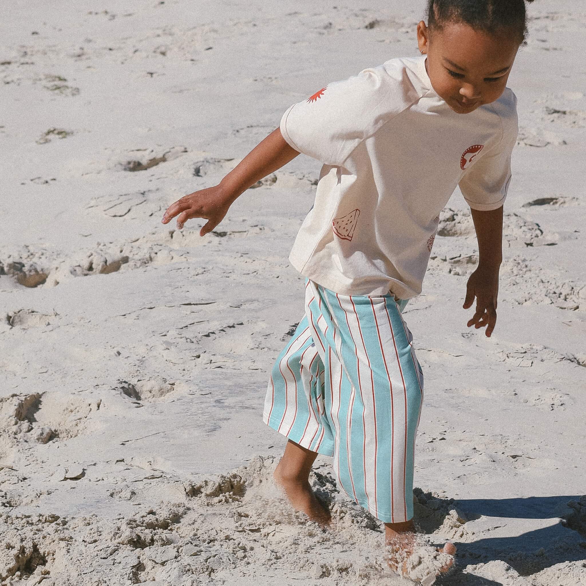 child wearing cream tee and blue and cream stripe pants on the beach