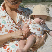 Baby in sun hat and printed onesie held by adult in matching shirt dress; sandy shoreline and blue sky.