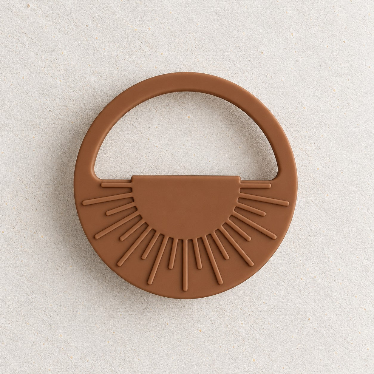 Clay silicone sun teether, a round teether with raised sunrays and an open handle, on a soft stone background